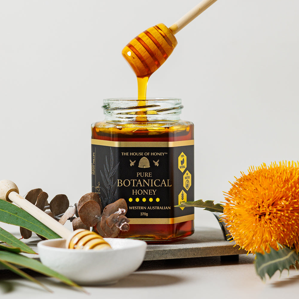 Jar of pure botanical honey with a honey dipper, surrounded by honeycomb and flowers on a light background.